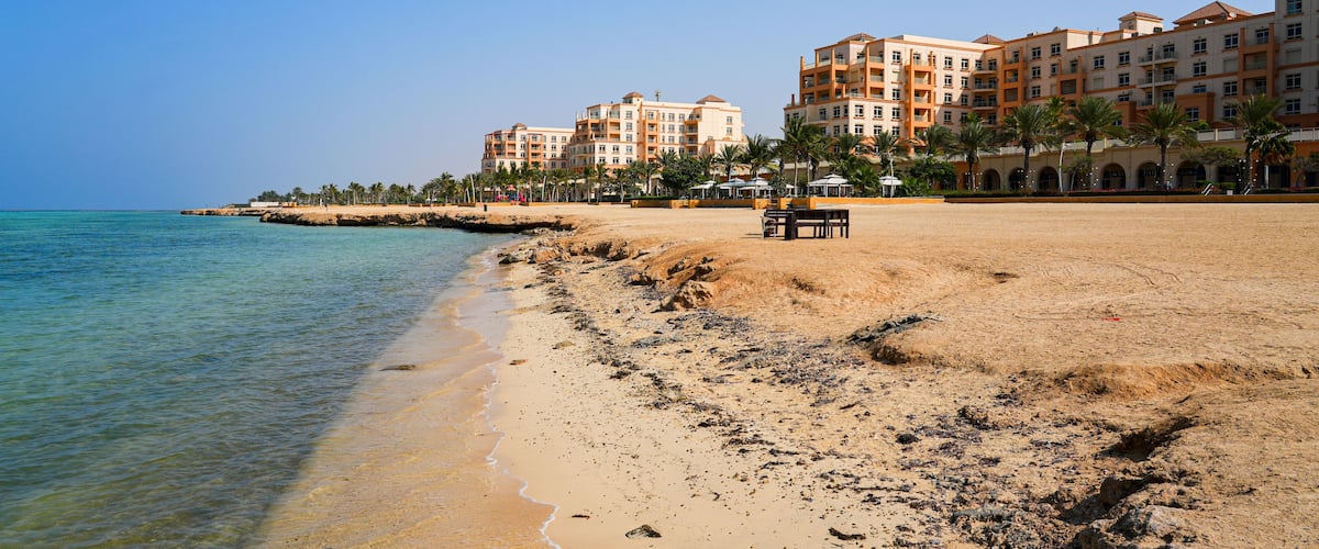 A sandy beach with clear water, palm trees, and a hotel in the background.