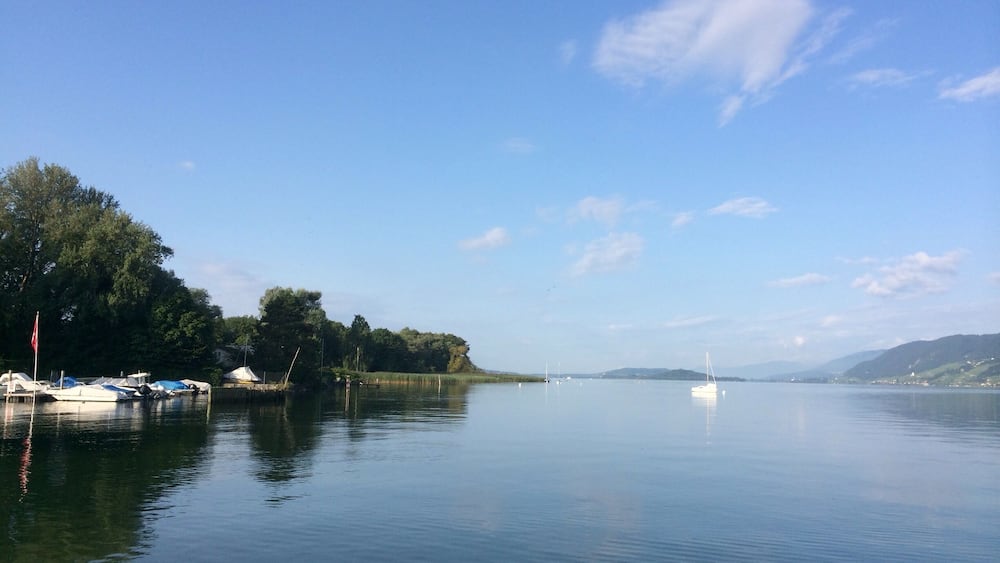 Ein ruhiger See mit anlegenden Booten links, umgeben von Bäumen und Hügeln im Hintergrund.
