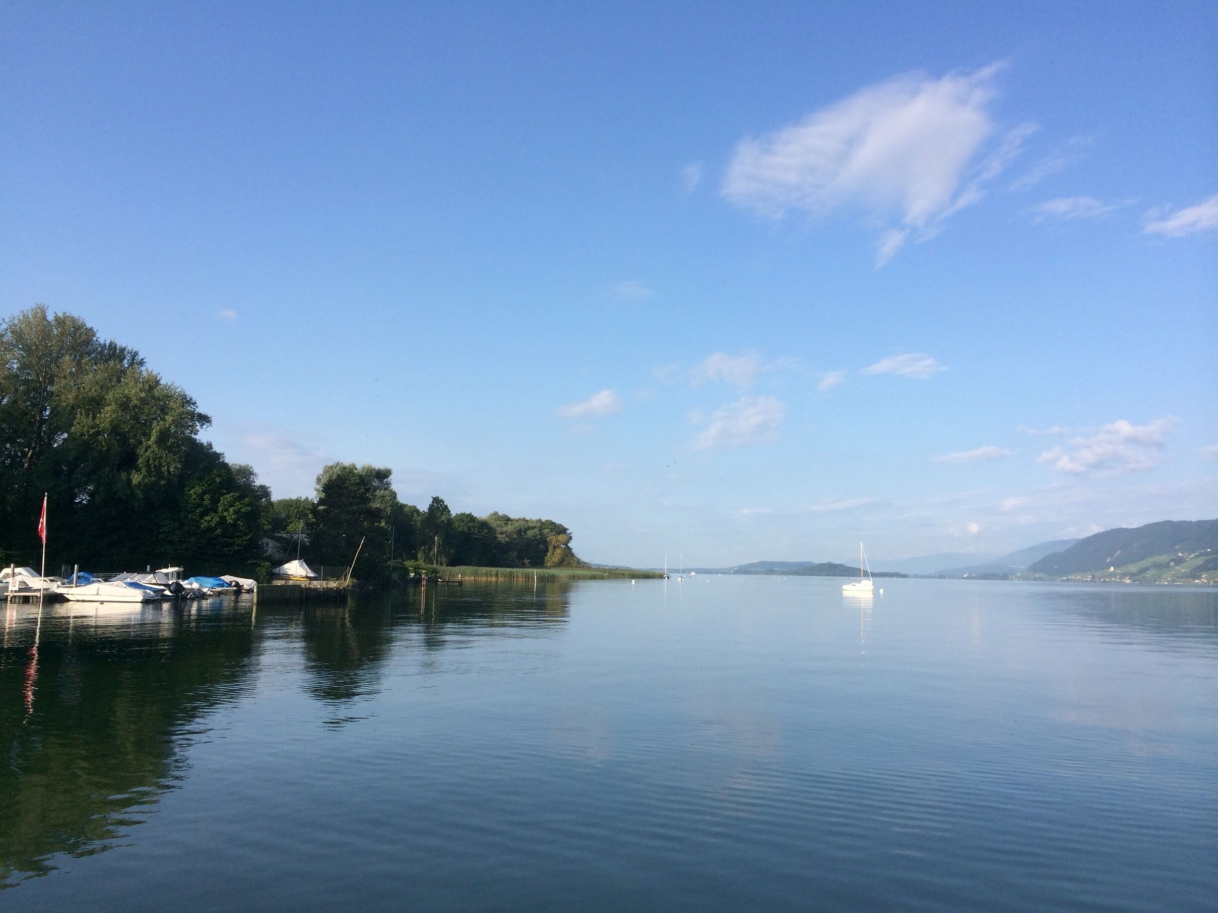 A calm lake with boats docked on the left, surrounded by trees and hills in the background.