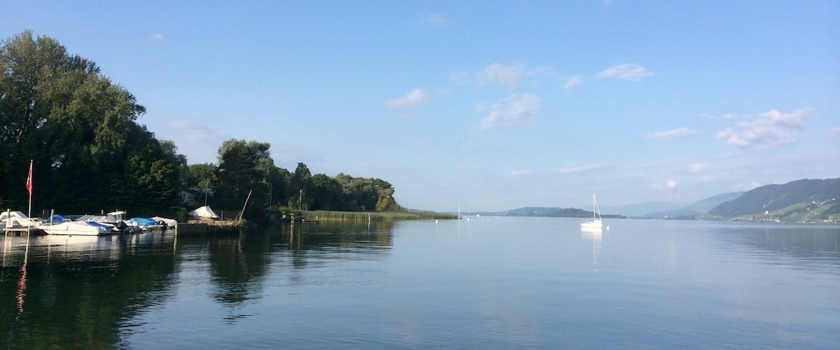 A calm lake with boats docked on the left, surrounded by trees and hills in the background.