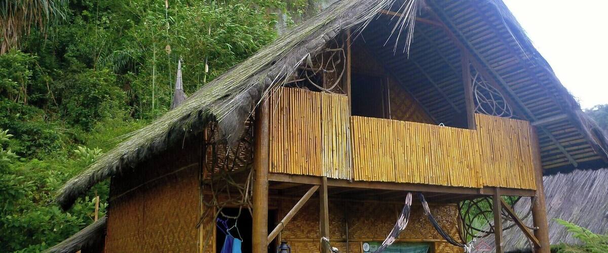A bamboo house with a thatched roof, situated in a lush green valley with a rocky cliff in the background.