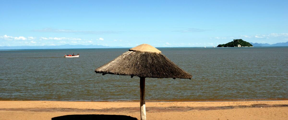 A beach with a thatched umbrella, a boat on the water, and a distant island.