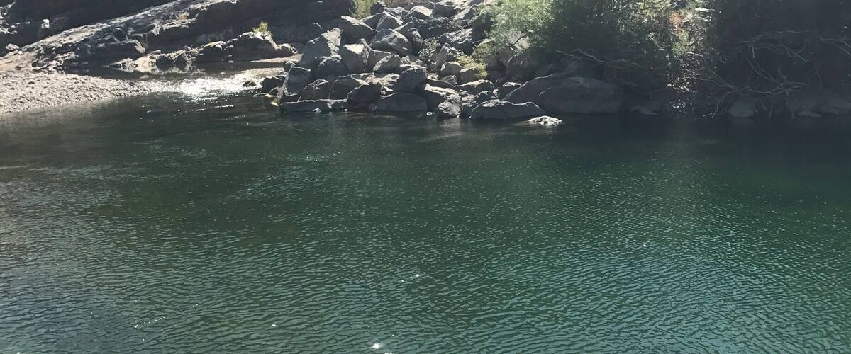 A clear river with sunlight reflecting on the water, surrounded by rocks and sparse vegetation.