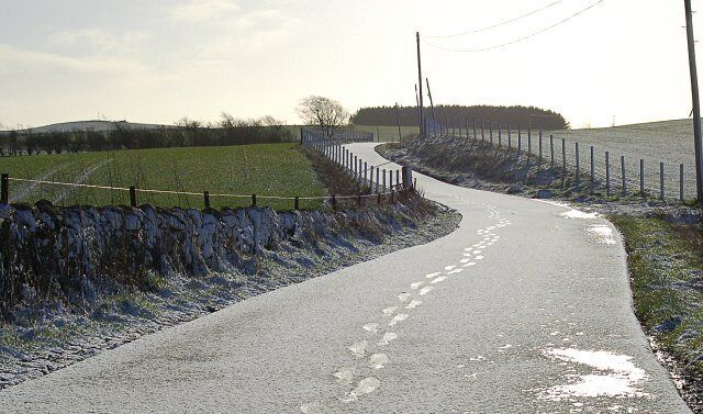 A rural road curving through a field with a stone wall on one side and a fence on the other.