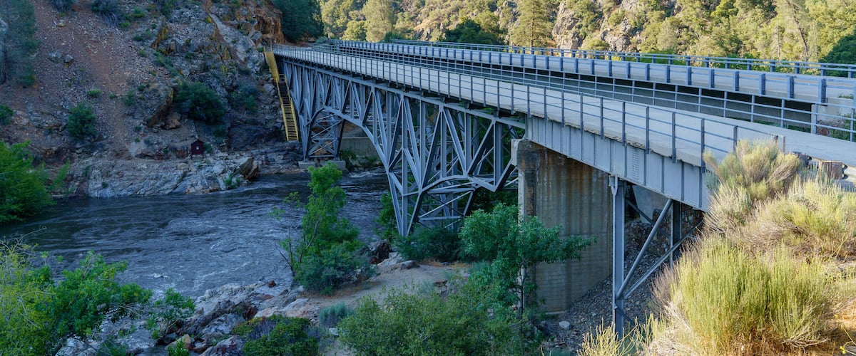 A steel bridge over a river with surrounding hills and greenery.