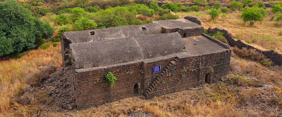 An ancient brick structure with a flat roof surrounded by dry grass and sparse trees in a hilly landscape.