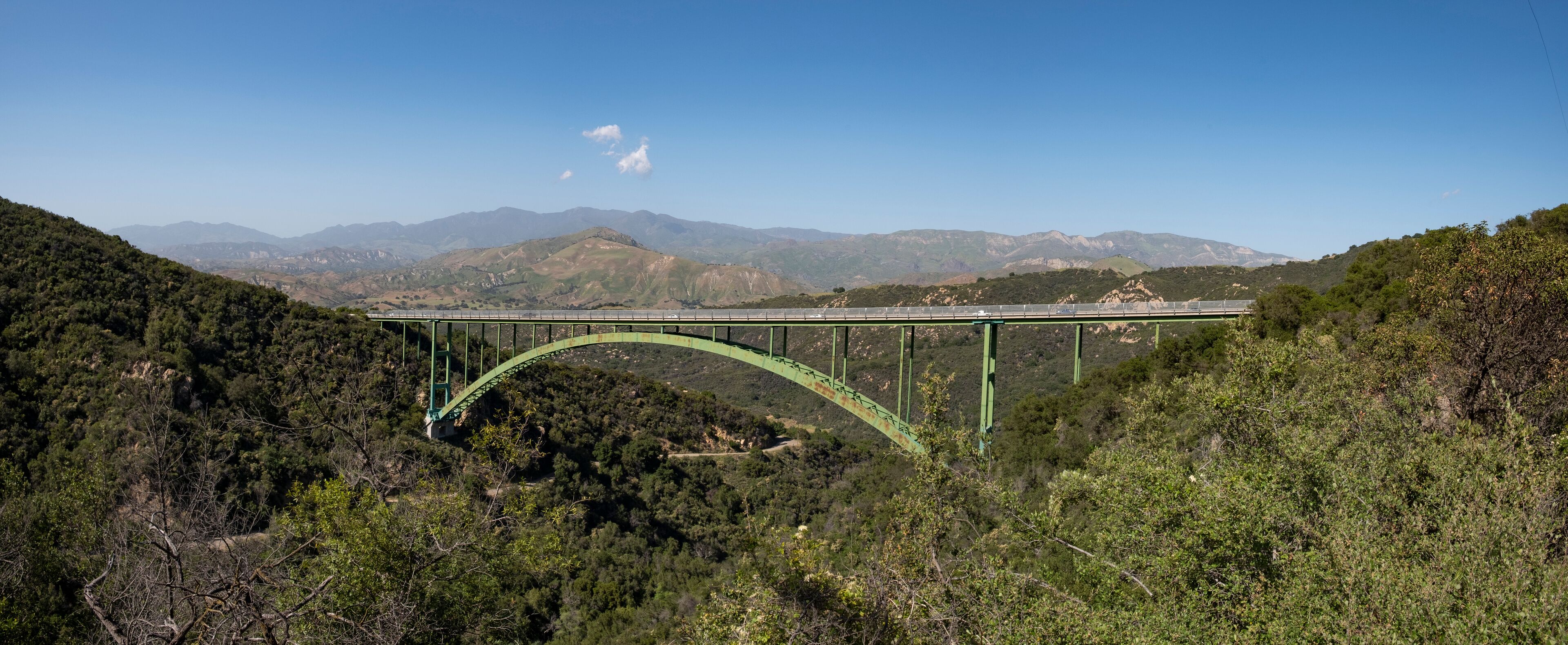 A green bridge over a valley with mountains in the background.