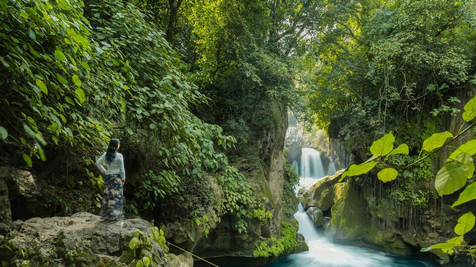 A person on a rope bridge over a turquoise river with a waterfall in the background.