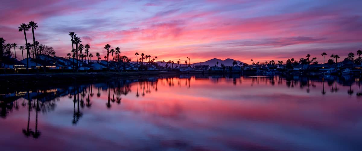 A sunset over a residential area with palm trees and houses reflecting on a calm body of water.