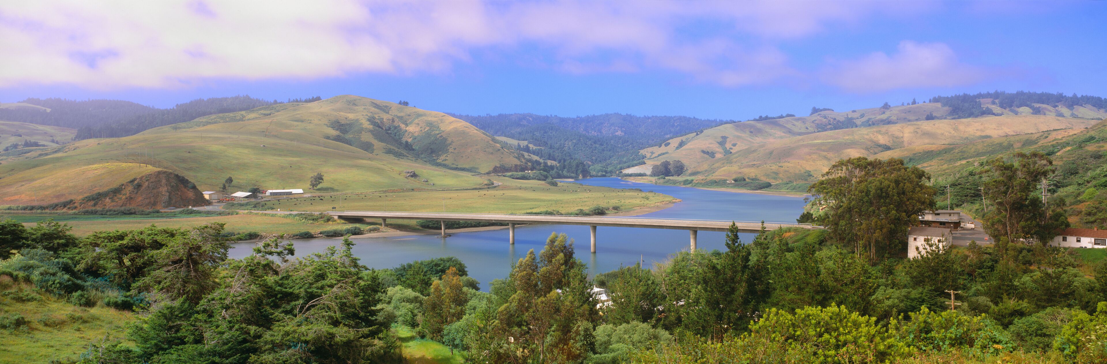 A river flowing through a valley with hills, a bridge, and a building near the water.