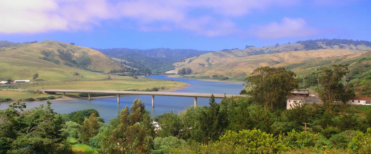 A river flowing through a valley with hills, a bridge, and a building near the water.