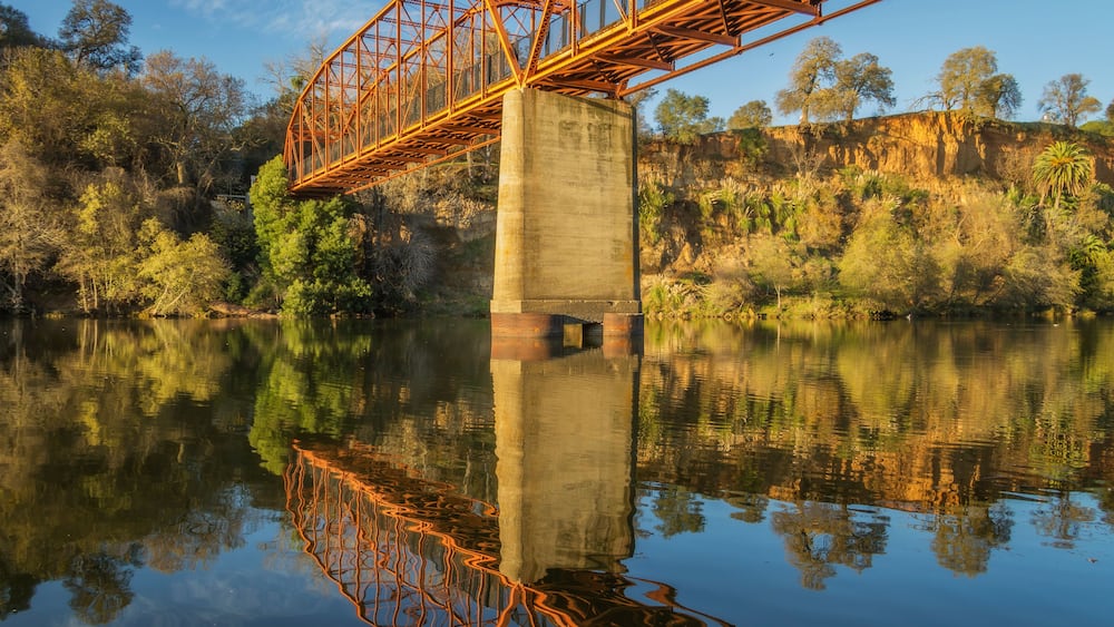 Eine Metallbrücke überspannt einen ruhigen Fluss, umgeben von Bäumen und Klippen.