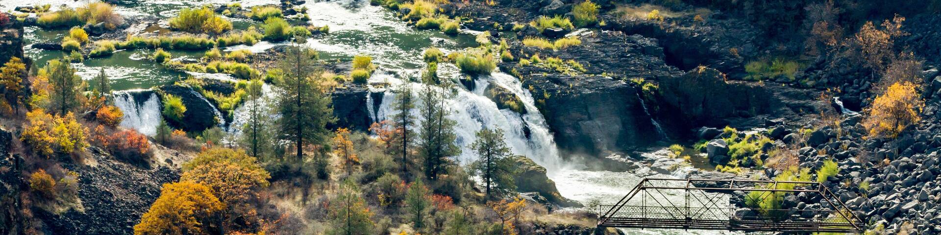 A river with waterfalls, surrounded by autumn-colored trees and a metal bridge.