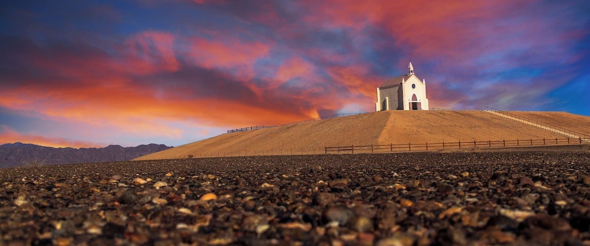 A small white church on a hill with a rocky foreground and a colorful sky.