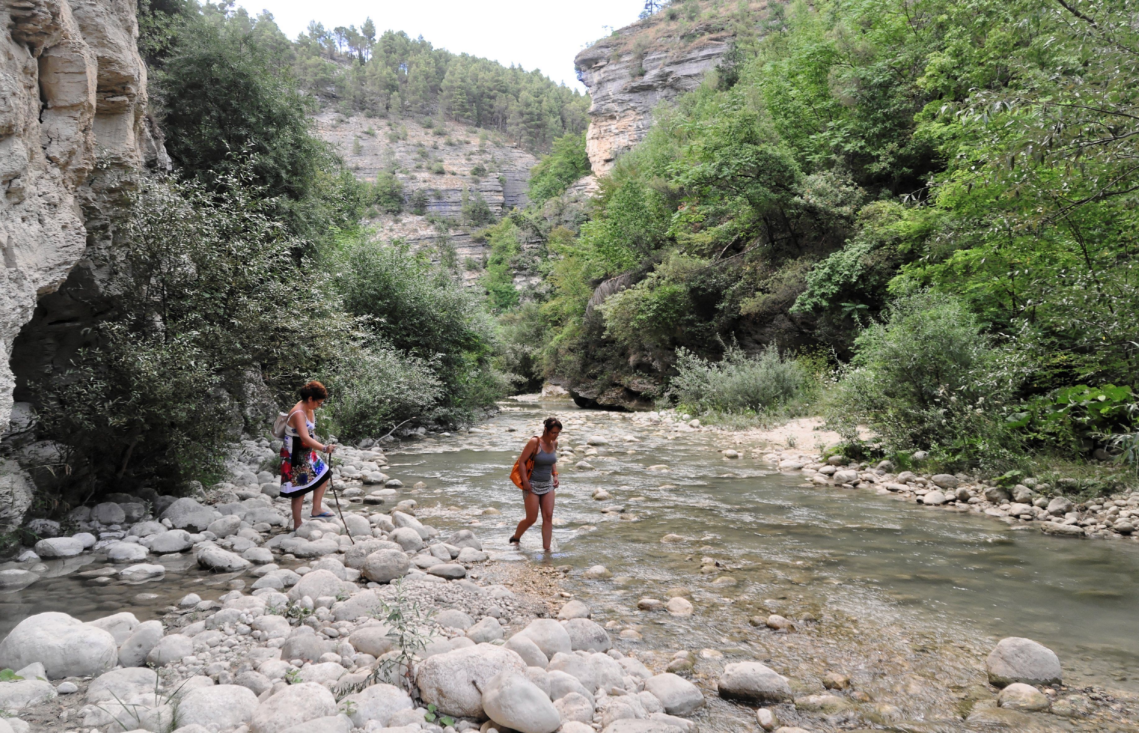 A rocky riverbed with two people standing in the water, surrounded by greenery and cliffs.