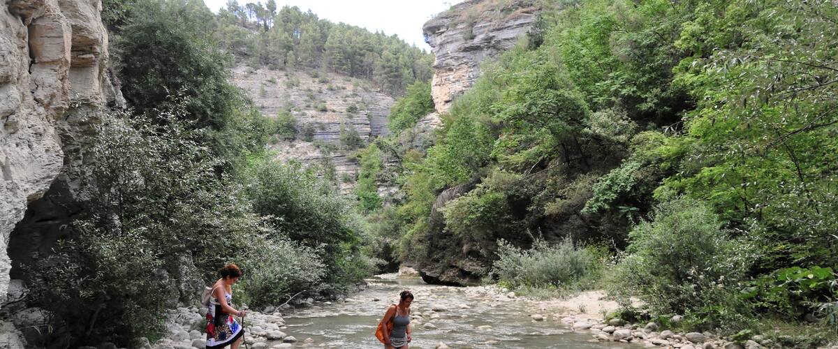 A rocky riverbed with two people standing in the water, surrounded by greenery and cliffs.