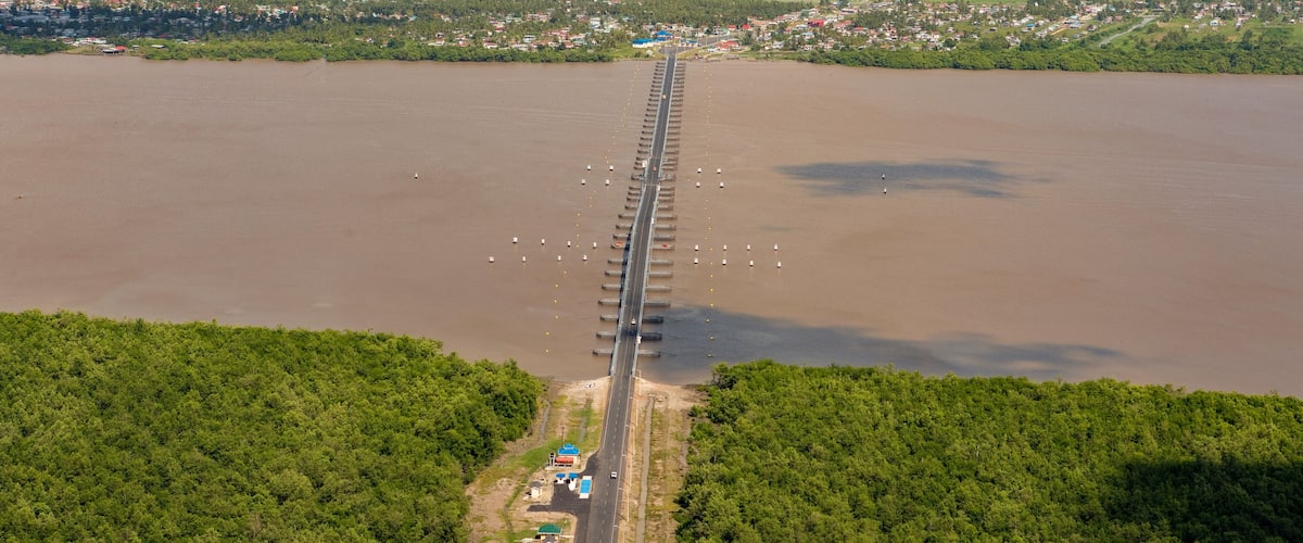 An aerial view of a long bridge over a wide river, surrounded by dense forests and a small town.