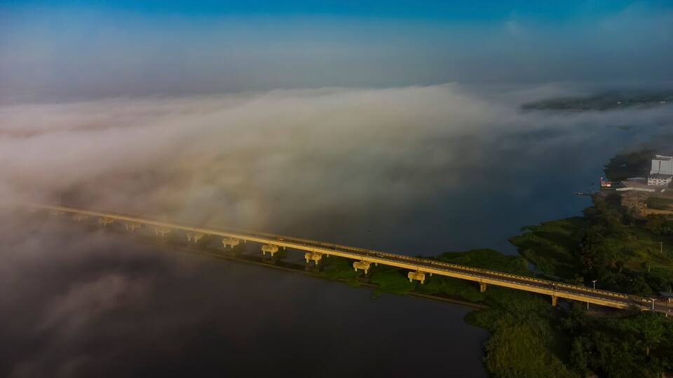A bridge over a body of water with fog and greenery on the sides.