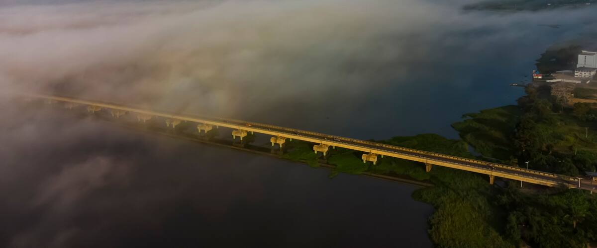 A bridge over a body of water with fog and greenery on the sides.