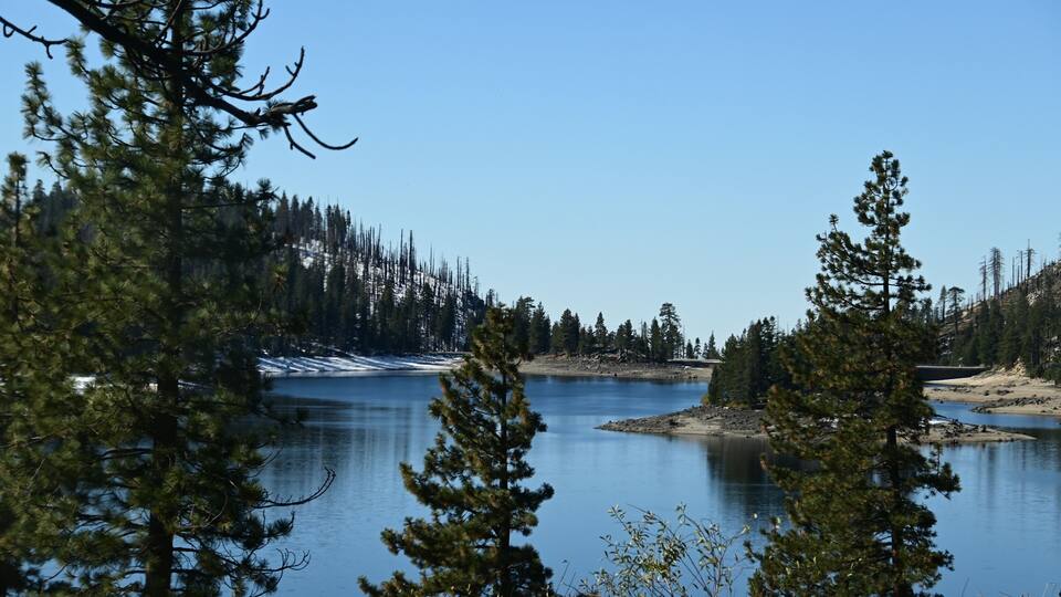 A calm lake surrounded by dense pine trees and snow patches.