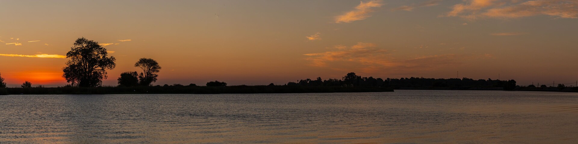 A sunset over a calm body of water with silhouettes of trees and a distant shoreline.