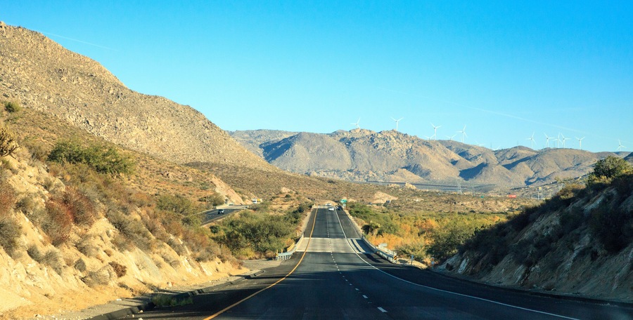 A highway with wind turbines in the distance, surrounded by mountainous terrain.
