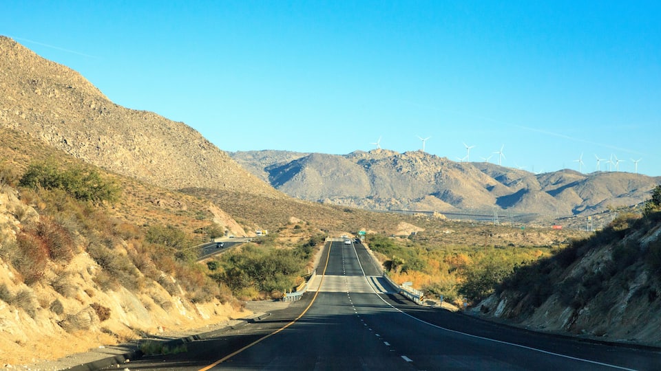 A highway with wind turbines in the distance, surrounded by mountainous terrain.