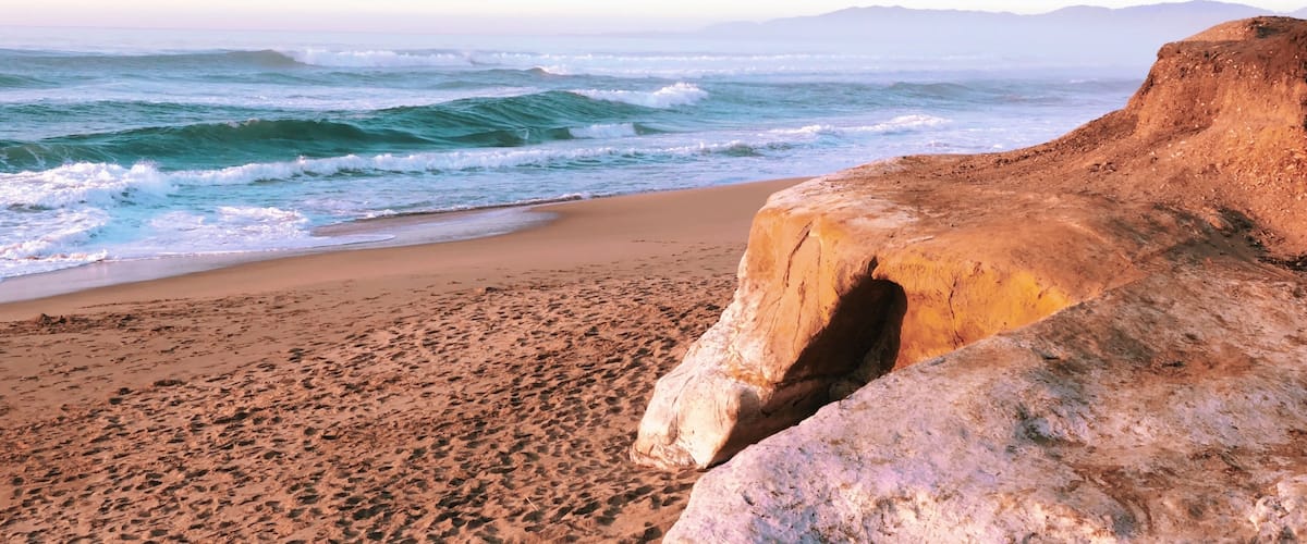 A beach with waves, a sandy shore, and a rocky outcrop.