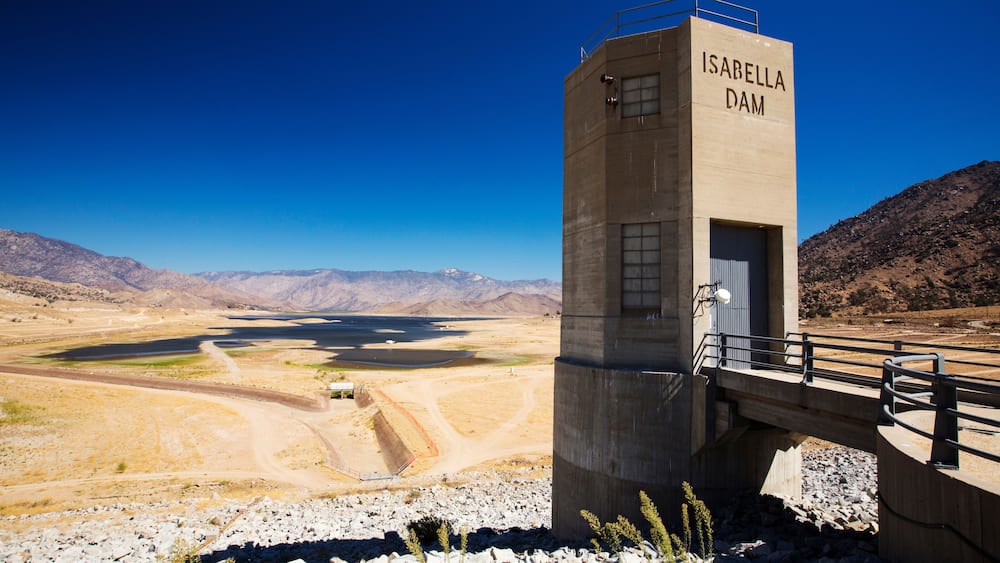 A concrete structure labeled 'ISABELLA DAM' with a bridge, a dry lakebed, and mountains in the background.