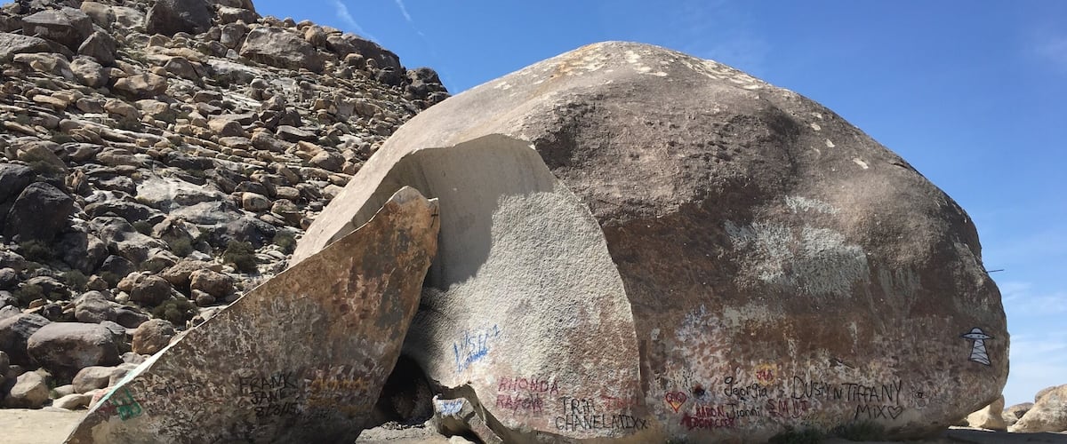 A large boulder with graffiti and a rocky hillside in the background.