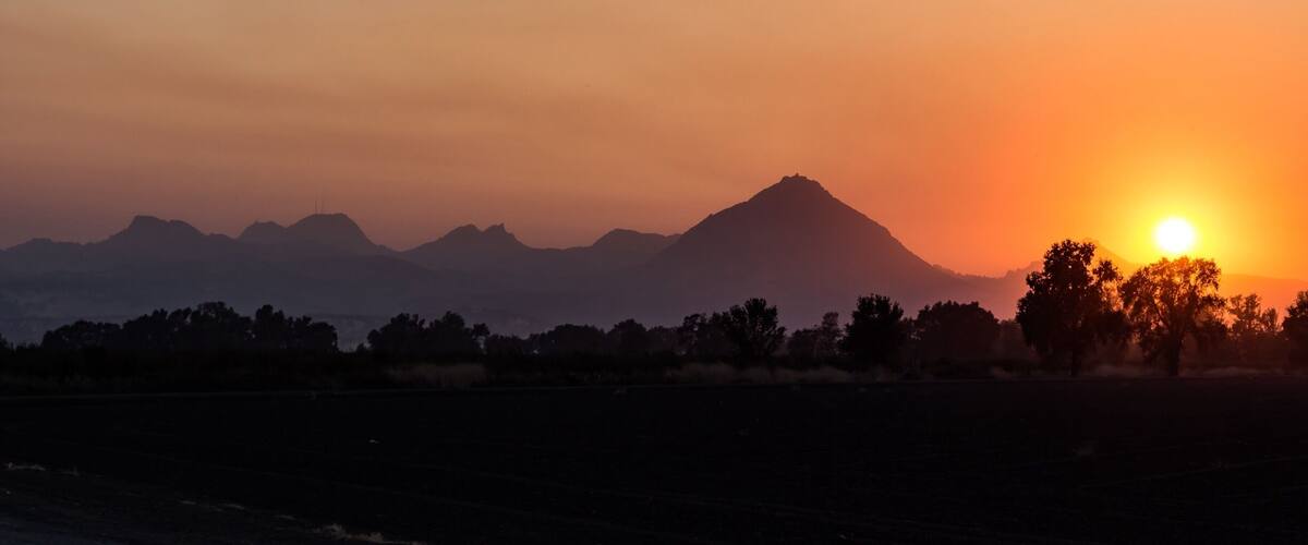 A sunset over a mountainous landscape with silhouettes of trees and a field.