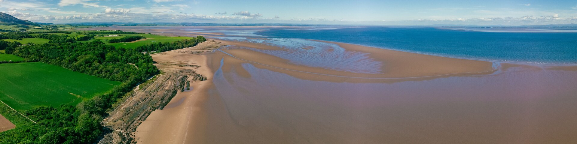 Un paisaje costero con una playa arenosa, campos verdes y un cielo azul despejado.