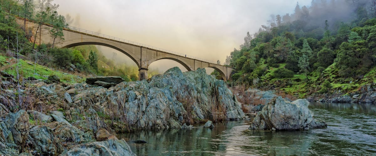 A stone bridge over a river with rocky banks and dense forest.