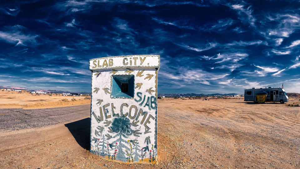 A weathered welcome sign in a desert landscape with a clear sky and a distant RV.