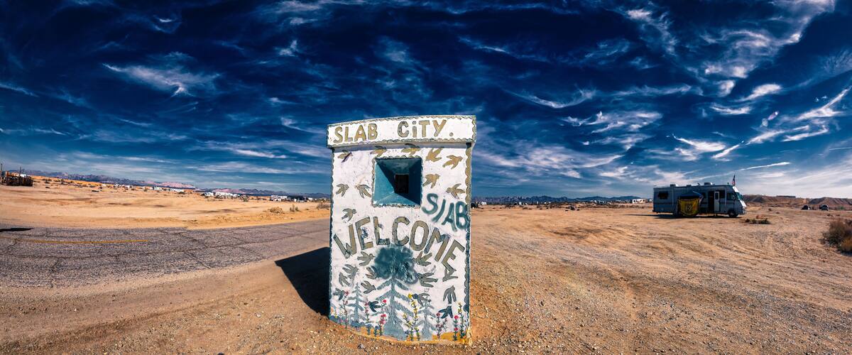 A weathered welcome sign in a desert landscape with a clear sky and a distant RV.