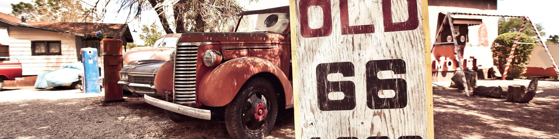 Un coche antiguo, un letrero de madera con la inscripción 'OLD 66 1926' y 'CALIFORNIA US 66', y un motel al fondo.