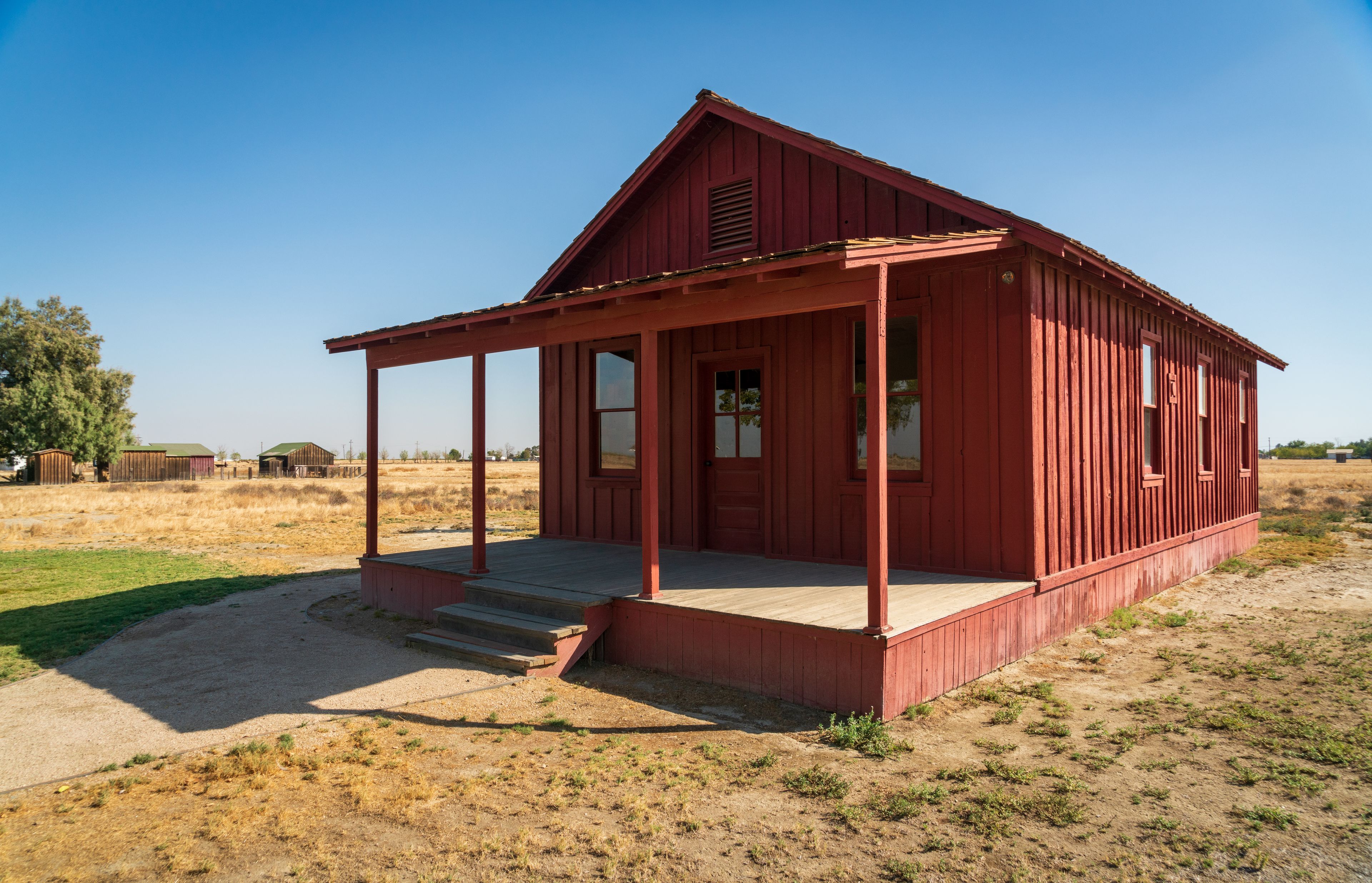 A red wooden building with a porch, surrounded by dry grass and a clear blue sky.