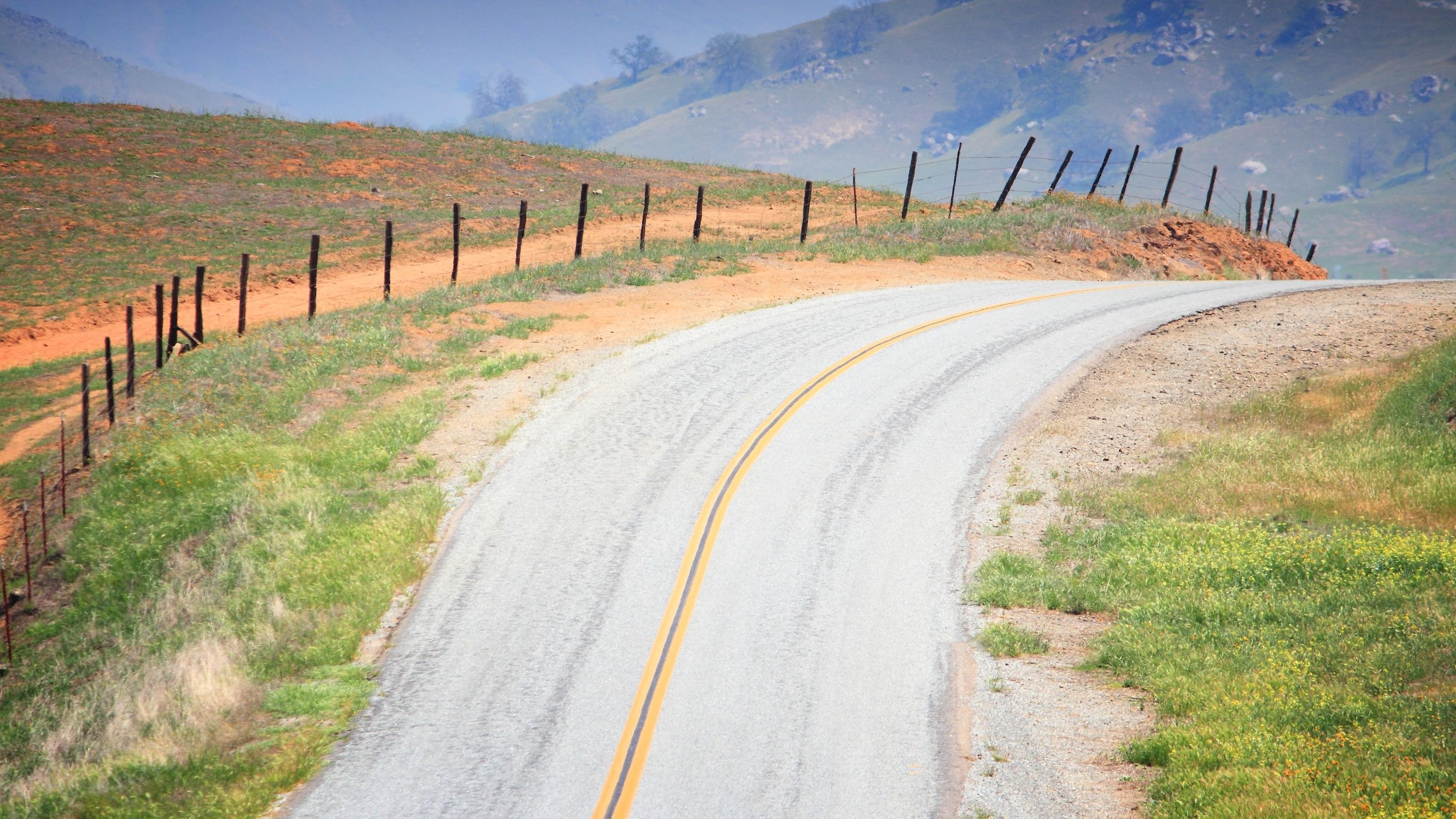 A winding road with a yellow centerline, bordered by a fence and grassy terrain, leading towards a hilly landscape.
