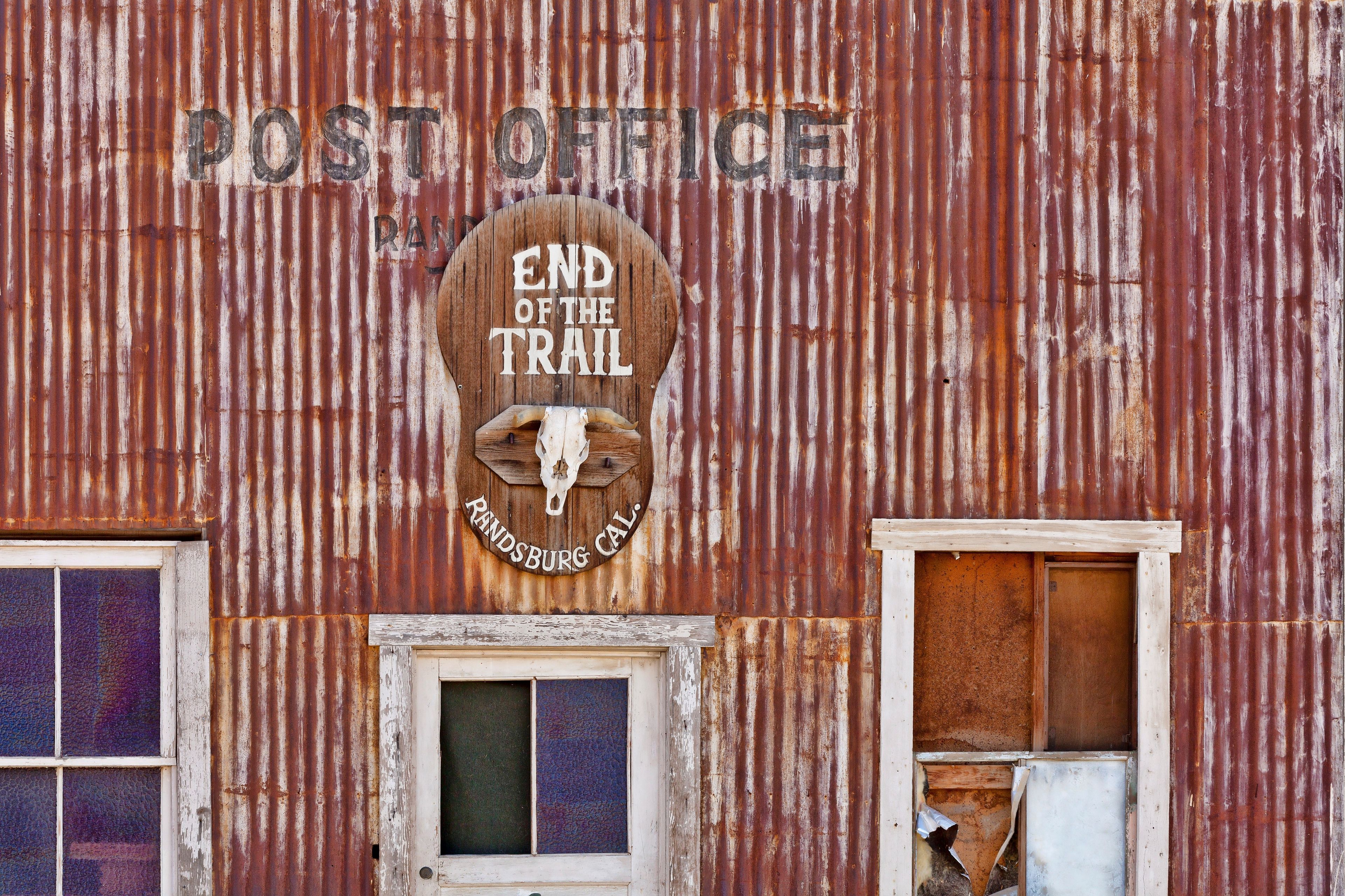 A rusted corrugated metal building with a sign that reads 'POST OFFICE END OF THE TRAIL RANDSBURG CAL.'