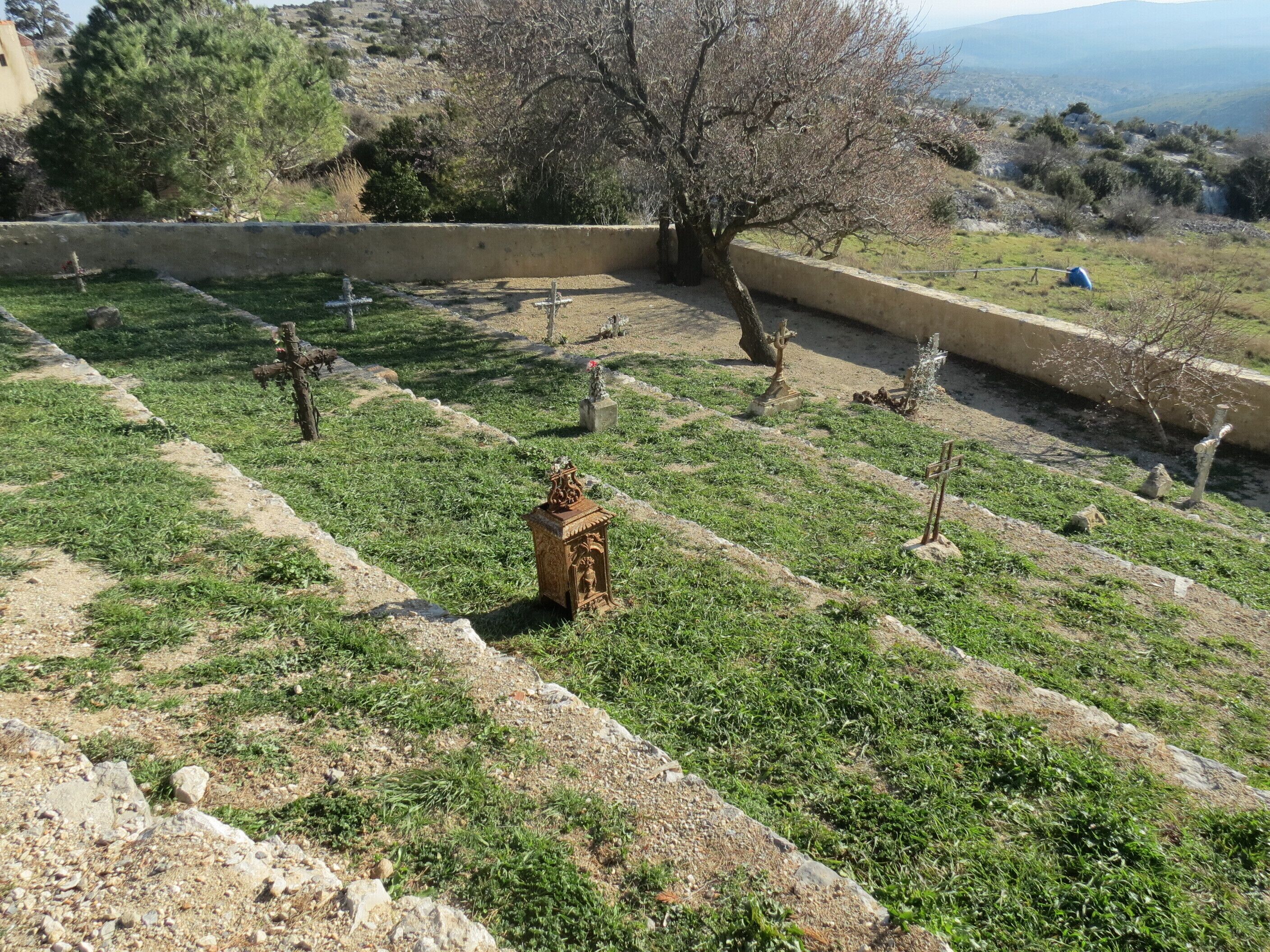 A cemetery with rows of graves, a central monument, and a backdrop of trees and hills.