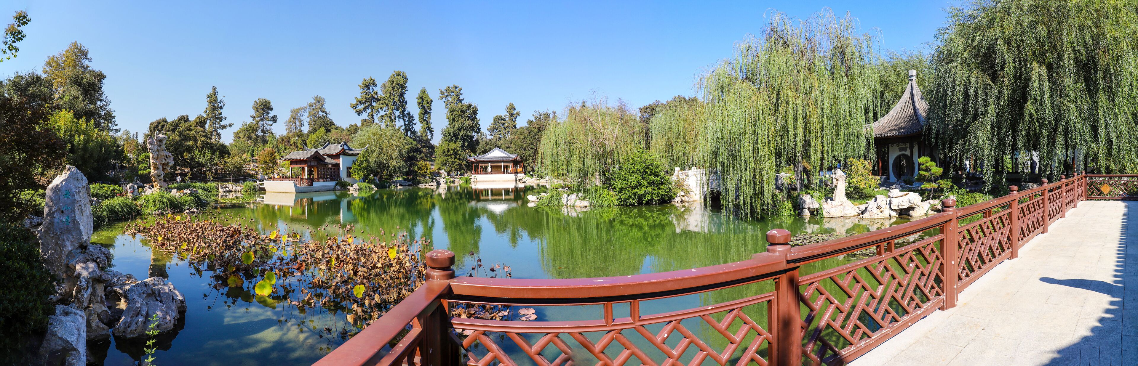 A serene pond with traditional Chinese architecture, including pagodas and a pavilion, surrounded by lush greenery and willow trees.