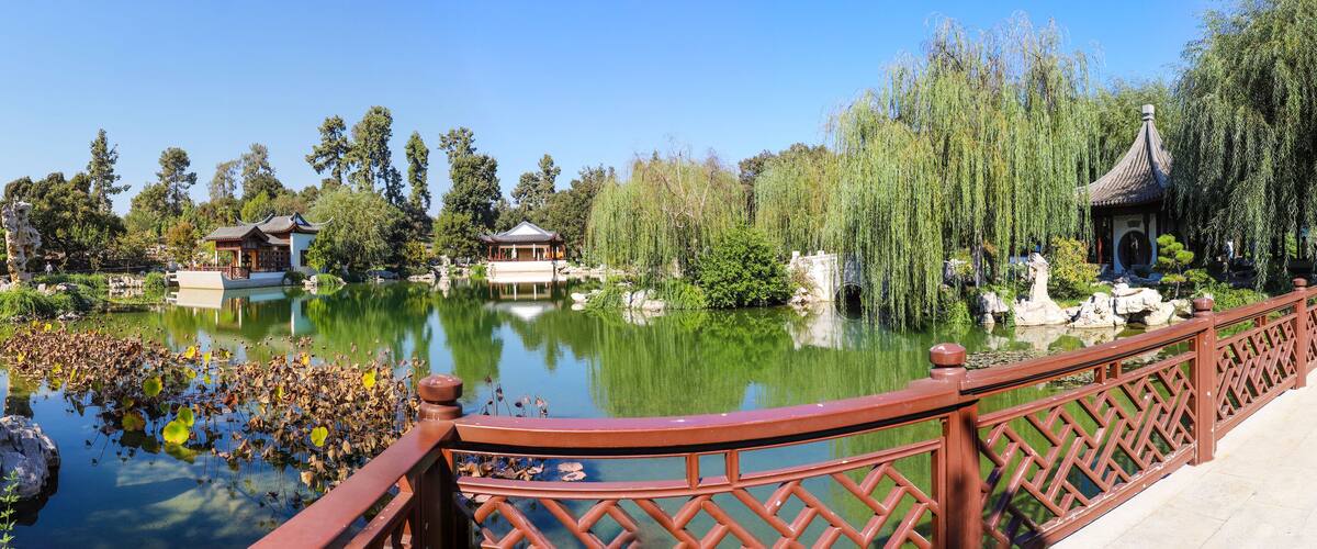 A serene pond with traditional Chinese architecture, including pagodas and a pavilion, surrounded by lush greenery and willow trees.