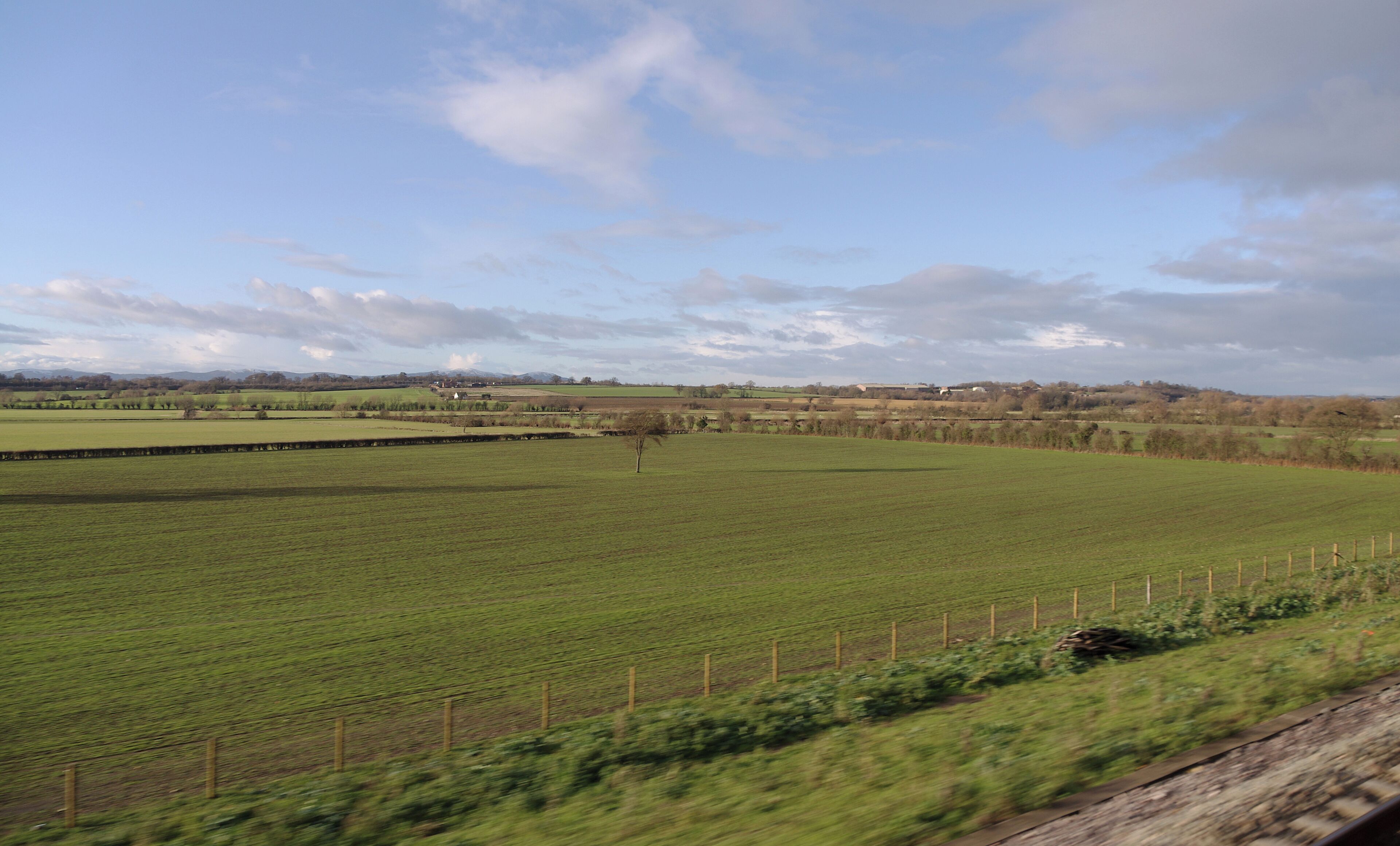 A rural landscape with green fields, a fence, and distant hills under a partly cloudy sky.