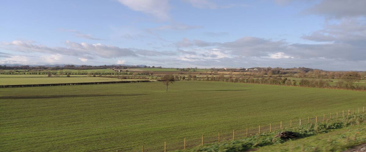 A rural landscape with green fields, a fence, and distant hills under a partly cloudy sky.