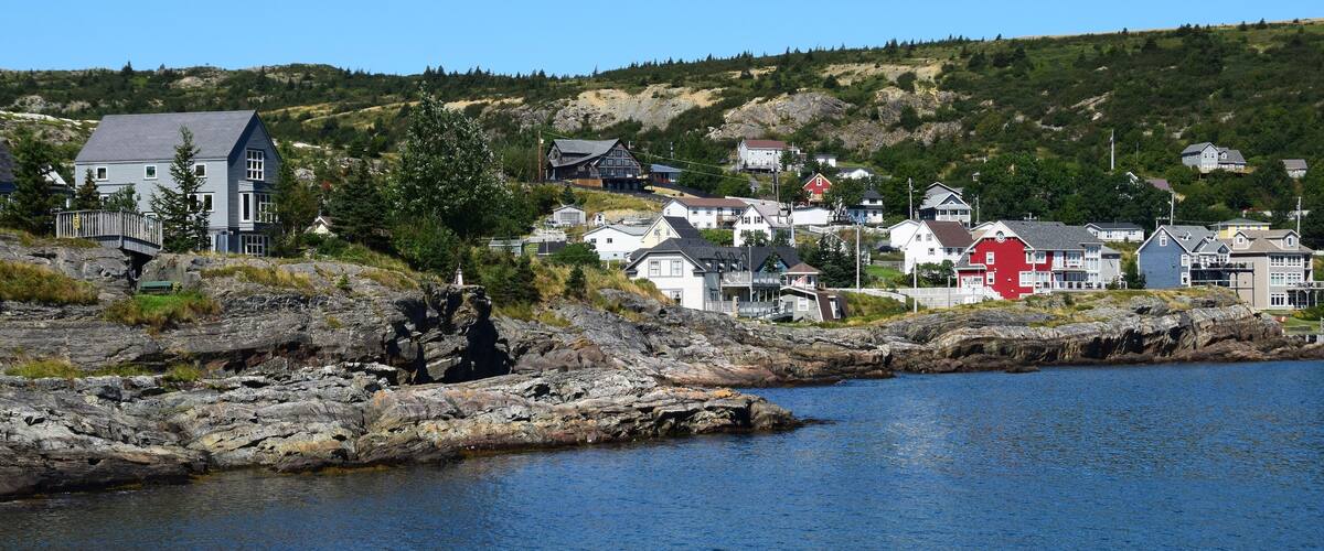 A coastal town with houses along the shoreline, a rocky outcrop, and a hill in the background.
