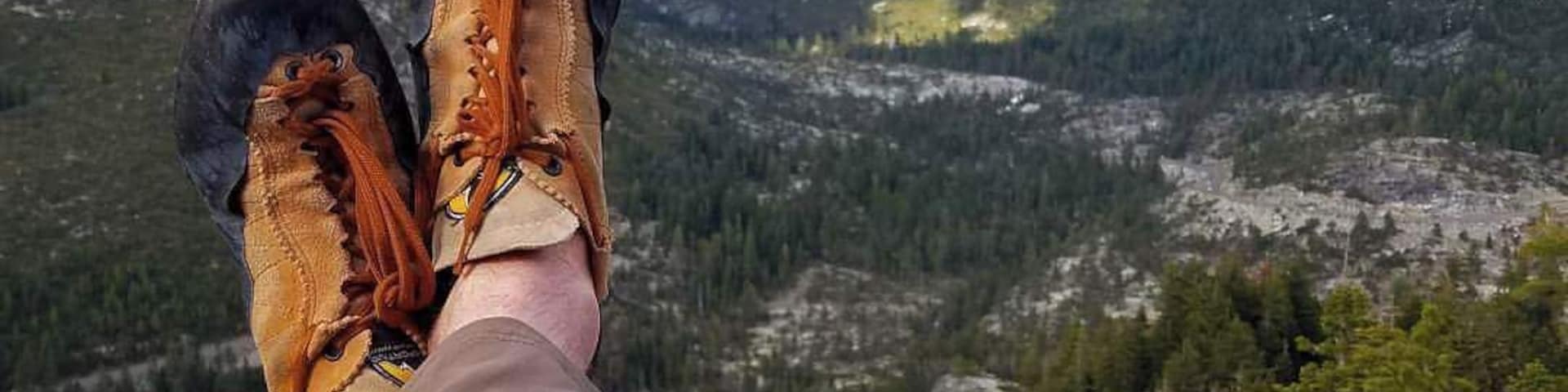 Piernas de una persona con botas de senderismo descansando sobre una roca, con vista a un paisaje montañoso de picos nevados y bosques densos.