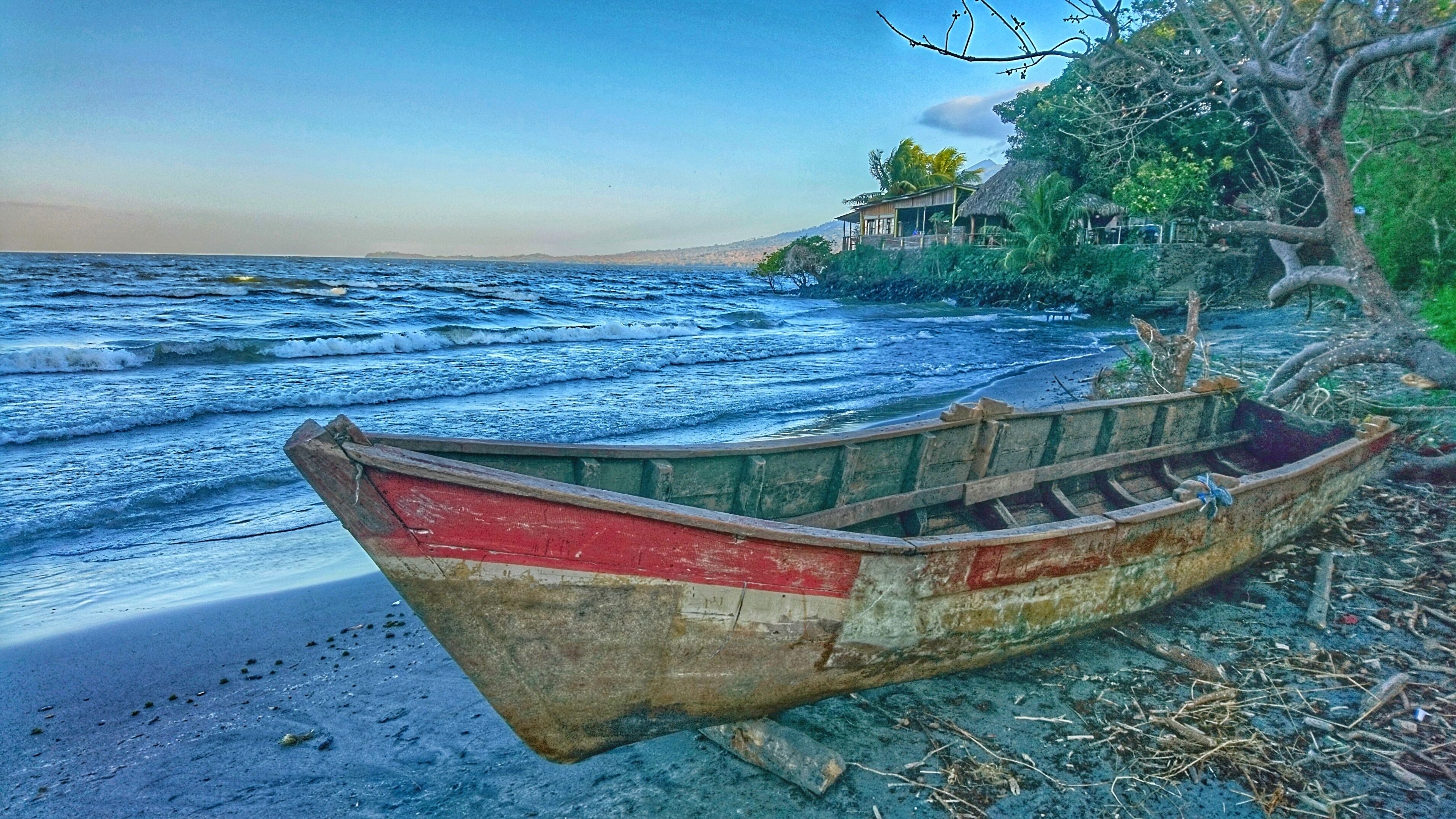 A weathered boat on a beach with waves, a house, and trees in the background.