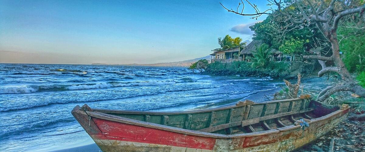 A weathered boat on a beach with waves, a house, and trees in the background.