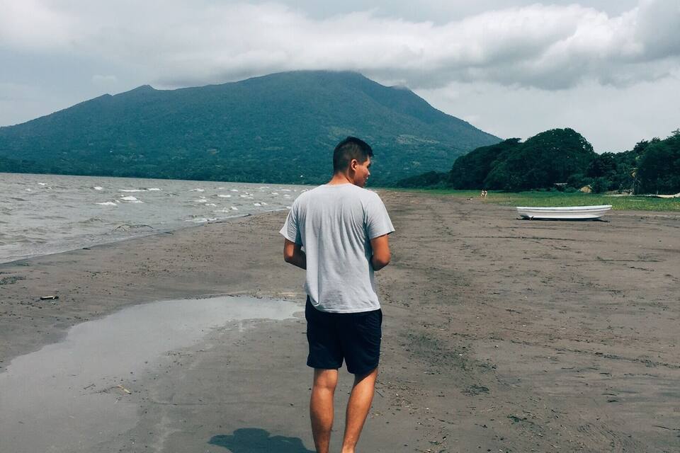Um homem está em pé numa praia, com um barco e uma montanha ao fundo.