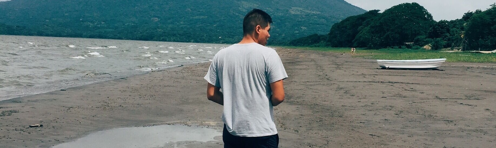 A man standing on a beach with a boat and a mountain in the background.